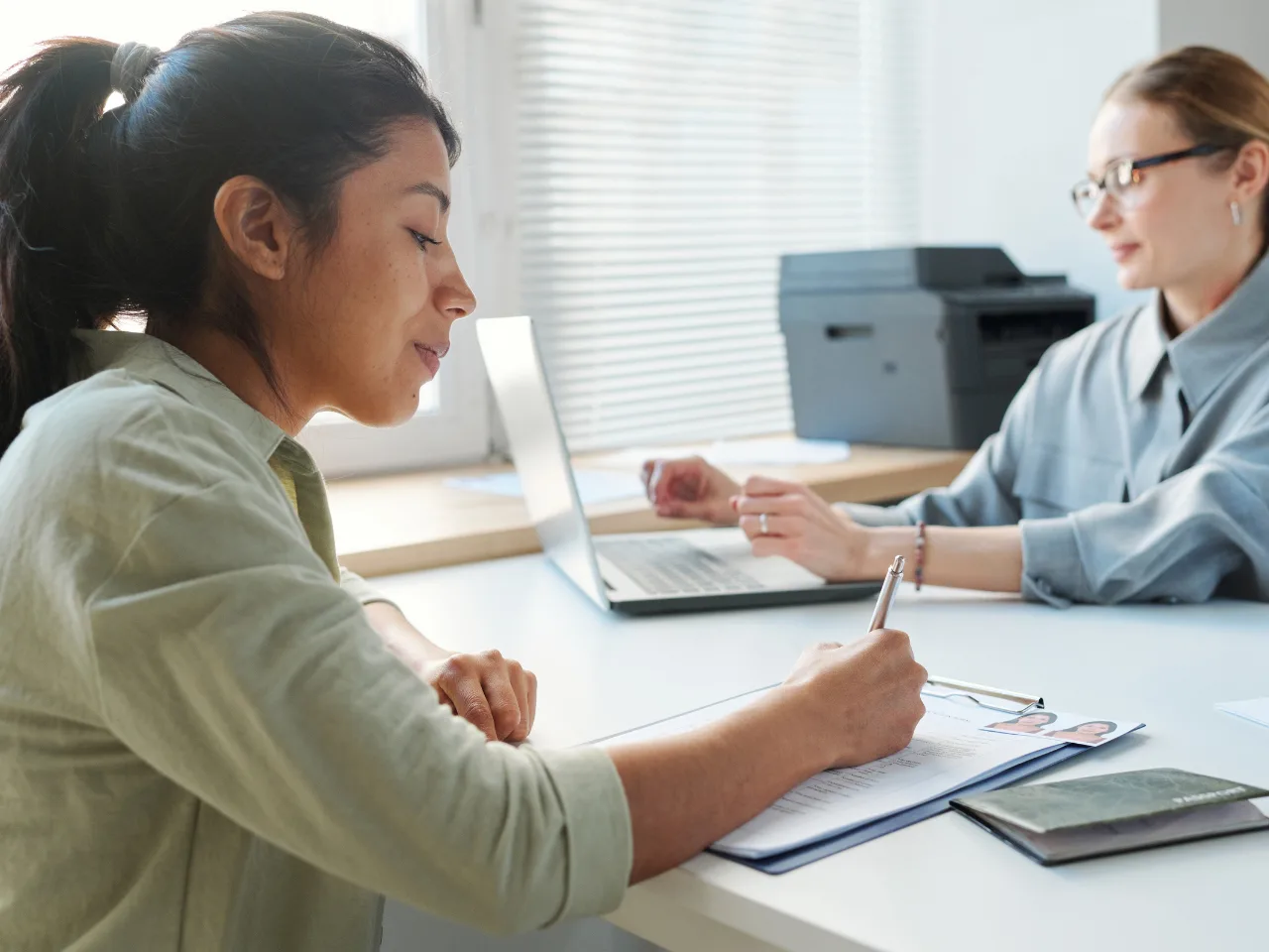 Two women sit at a desk in a bright office; one is writing on a form with a pen, while the other works on a laptop. A printer and documents are on the desk, and natural light comes through the window blinds.