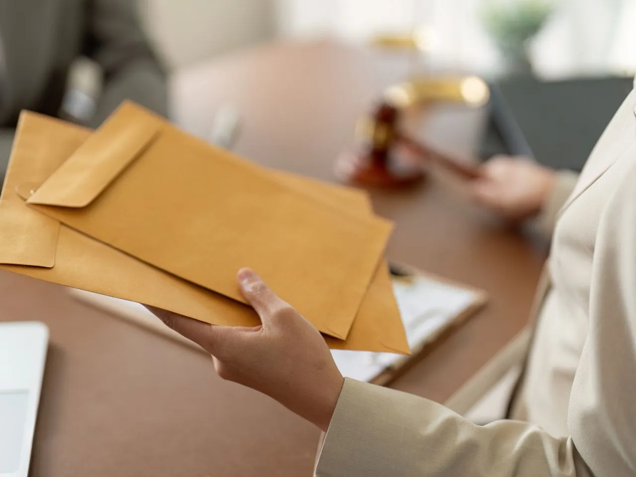 A person in a suit holding several large brown envelopes. In the background, a blurred out desk is visible with a gavel and paperwork on it. The scene appears to be in an office or professional setting.