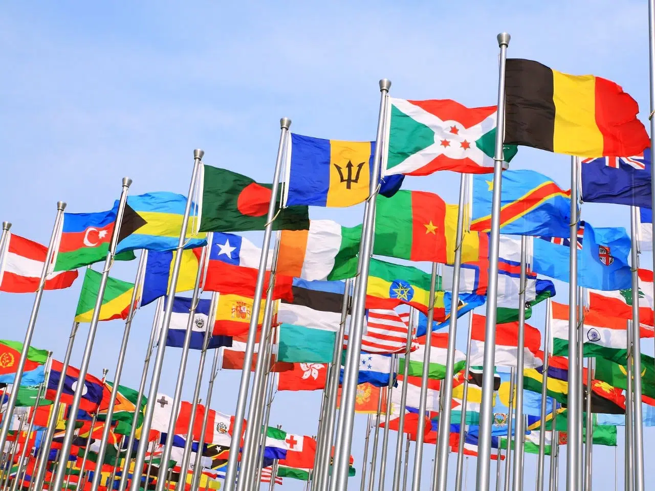 Numerous international flags on tall flagpoles against a clear blue sky, displaying various colors and patterns from different countries. The flags are fluttering in the wind, symbolizing global unity and diversity.