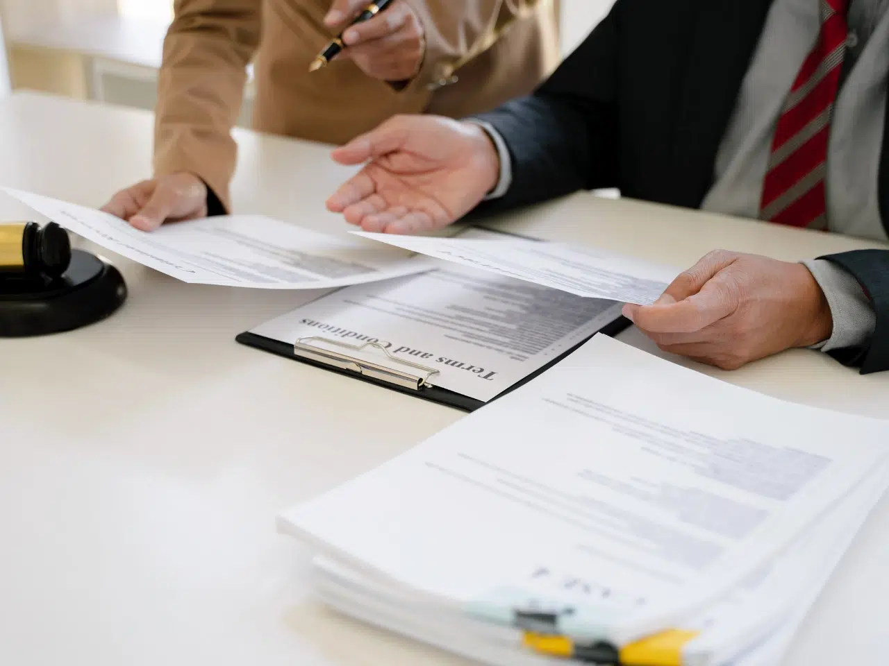 Two people in business attire review documents at a desk covered with paperwork, a clipboard labeled Terms and Conditions, and a gavel visible in the corner, suggesting a legal or contract discussion.