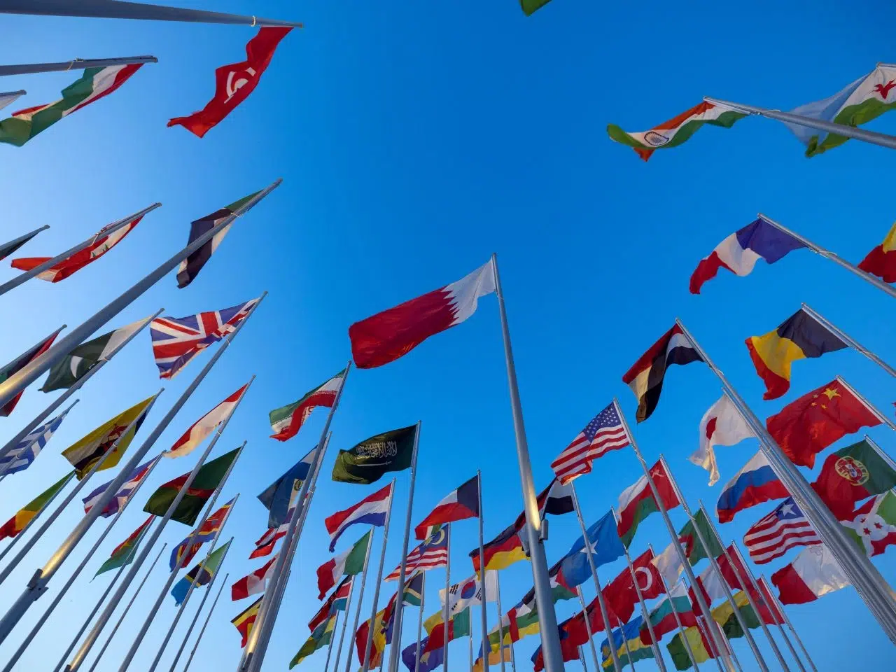 Multiple international flags on tall poles waving against a clear blue sky, viewed from below. The flags represent various countries with distinct colors and patterns, creating a vibrant and multicultural scene.