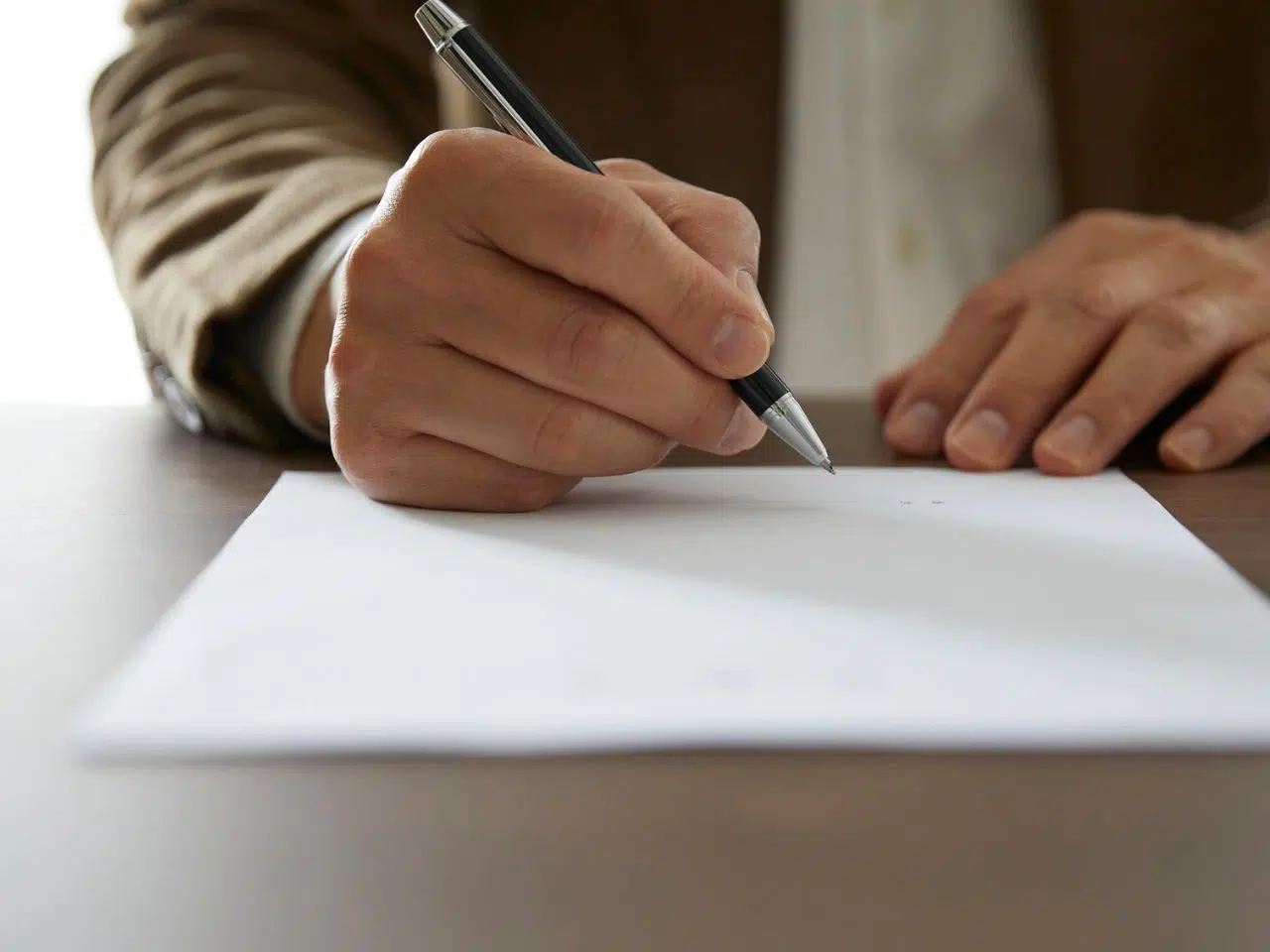 A person in a brown suit is writing on a blank sheet of paper with a black pen. The focus is on the hands, with the paper resting on a wooden table.