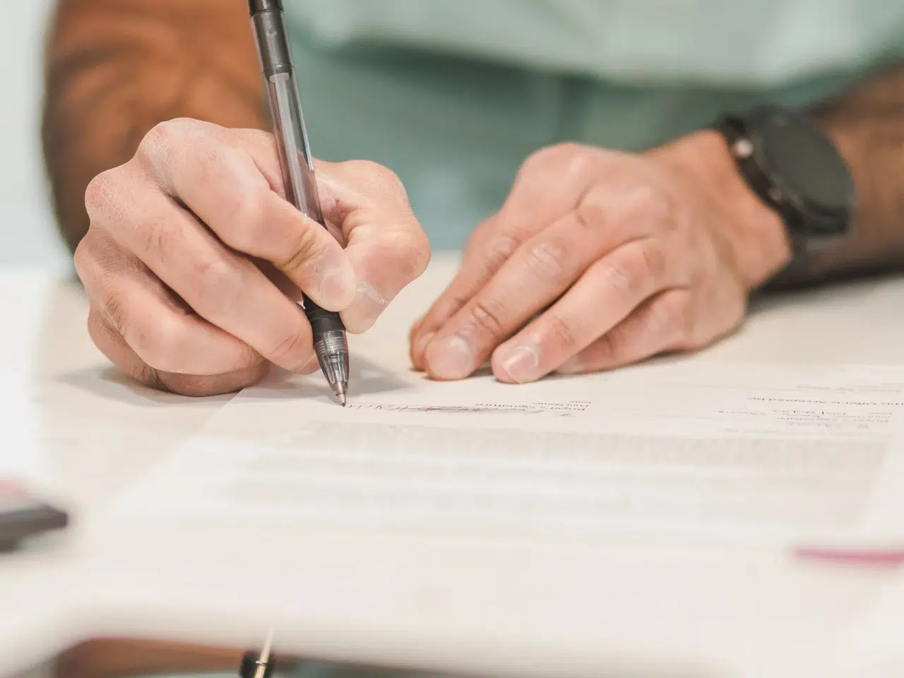 A person wearing a watch is holding a pen and signing or writing on a document placed on a white table. The focus is on the hands and the paper.