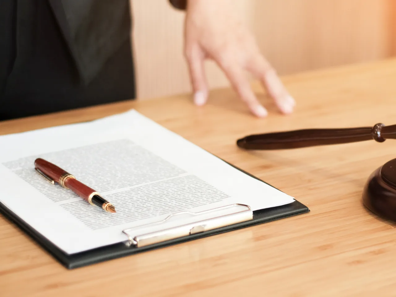 A close-up of a clipboard with a document and a pen on a wooden desk, next to a gavel and a persons hand resting on the table.