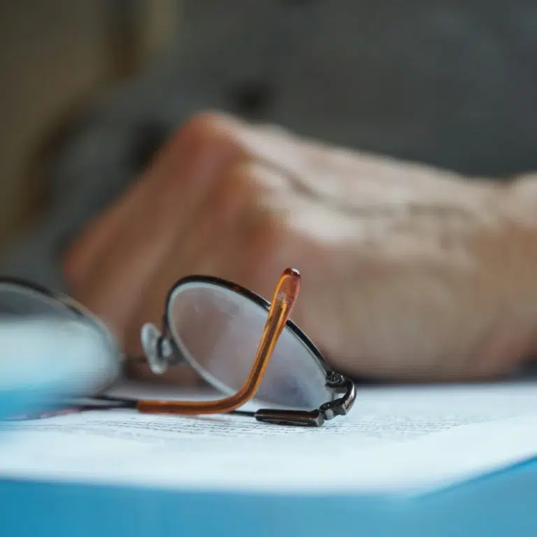 Close-up of a pair of reading glasses resting on an open book page with text. A hand is visible in the background, slightly out of focus, suggesting someone is reading or preparing to pick up the glasses.
