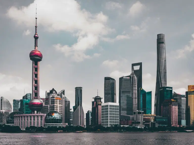Skyline of a modern city featuring diverse skyscrapers, including a prominent tower with a spherical observation deck and a unique building with a square-shaped opening at the top, under a cloudy sky.