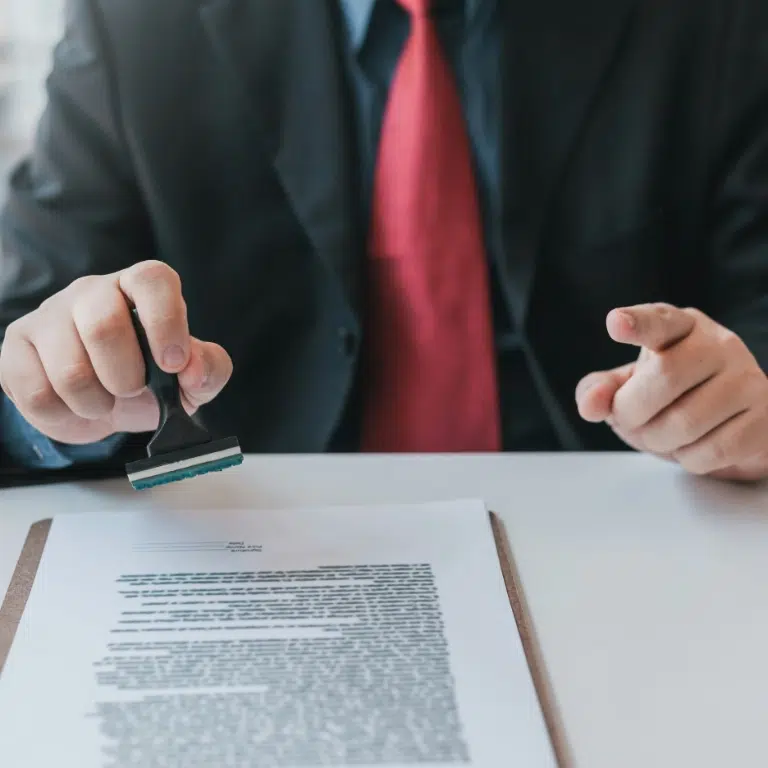 A person in a suit holds a rectangular stamp over a document on a desk. The document is placed on a clipboard. The persons other hand is partially visible, pointing at the document. The background is blurred.
