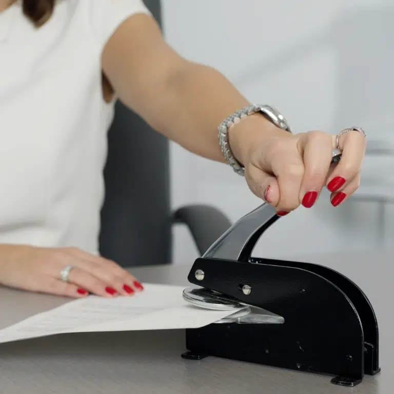 A person with red nail polish uses a black metal embosser on a sheet of paper. They are wearing a white shirt, a bracelet, and a watch. The scene is set against a neutral background.