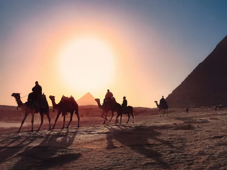 Silhouetted against a setting sun, riders on camels traverse the desert near the pyramids. The sky is a gradient of warm hues, and the ground and pyramids cast long shadows, creating a dramatic and timeless scene.