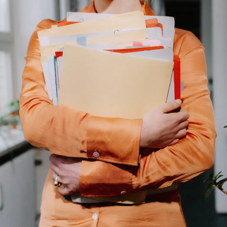 A person wearing an orange shirt holds a stack of assorted folders and documents. The scene appears to be in a bright indoor setting with natural light. Several windows are blurred in the background.
