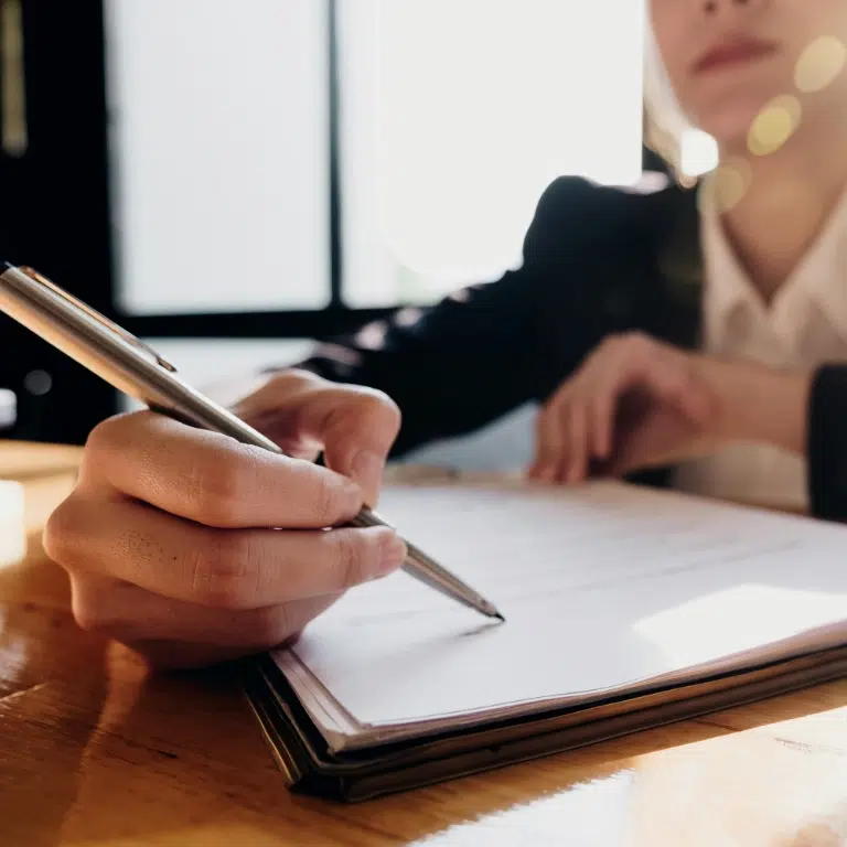A person in a suit is sitting at a table, holding a silver pen and signing or writing on a document. The background is softly lit, creating a focused atmosphere on the action.
