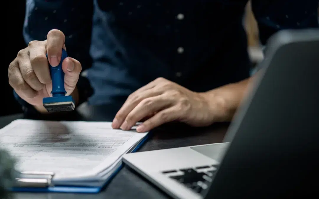 A person in a dark shirt presses a blue stamp onto a document on a clipboard beside an open laptop, suggesting approval or verification of paperwork in an office setting.