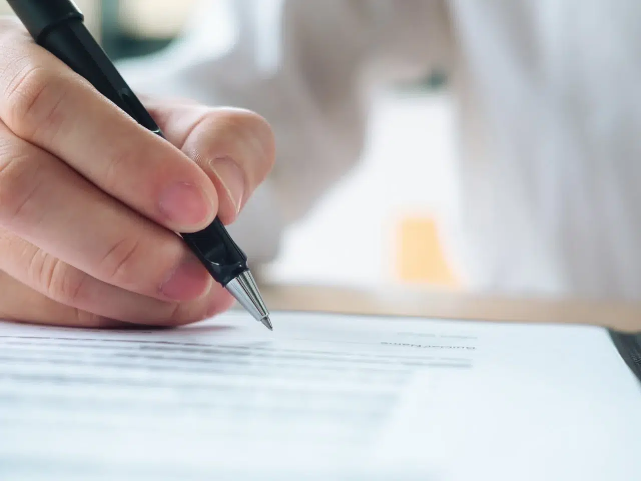 A close-up of a hand holding a pen and signing or writing on a document with printed text. The background is out of focus, drawing attention to the writing action.