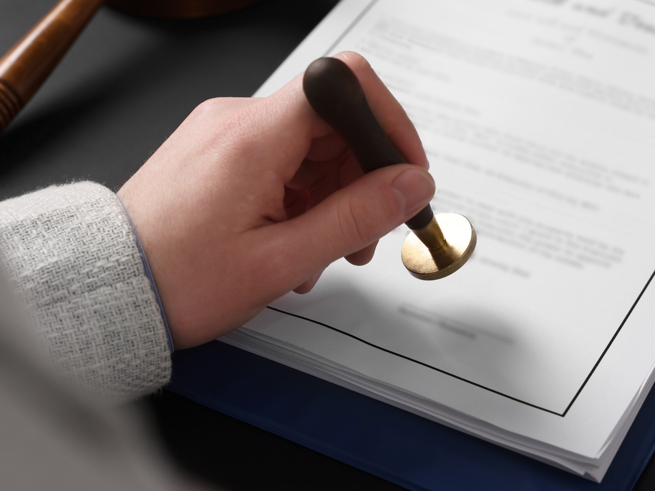 A person presses a round stamp onto a stack of official-looking documents, signifying approval or validation. A gavel is partially visible in the background on a dark surface.