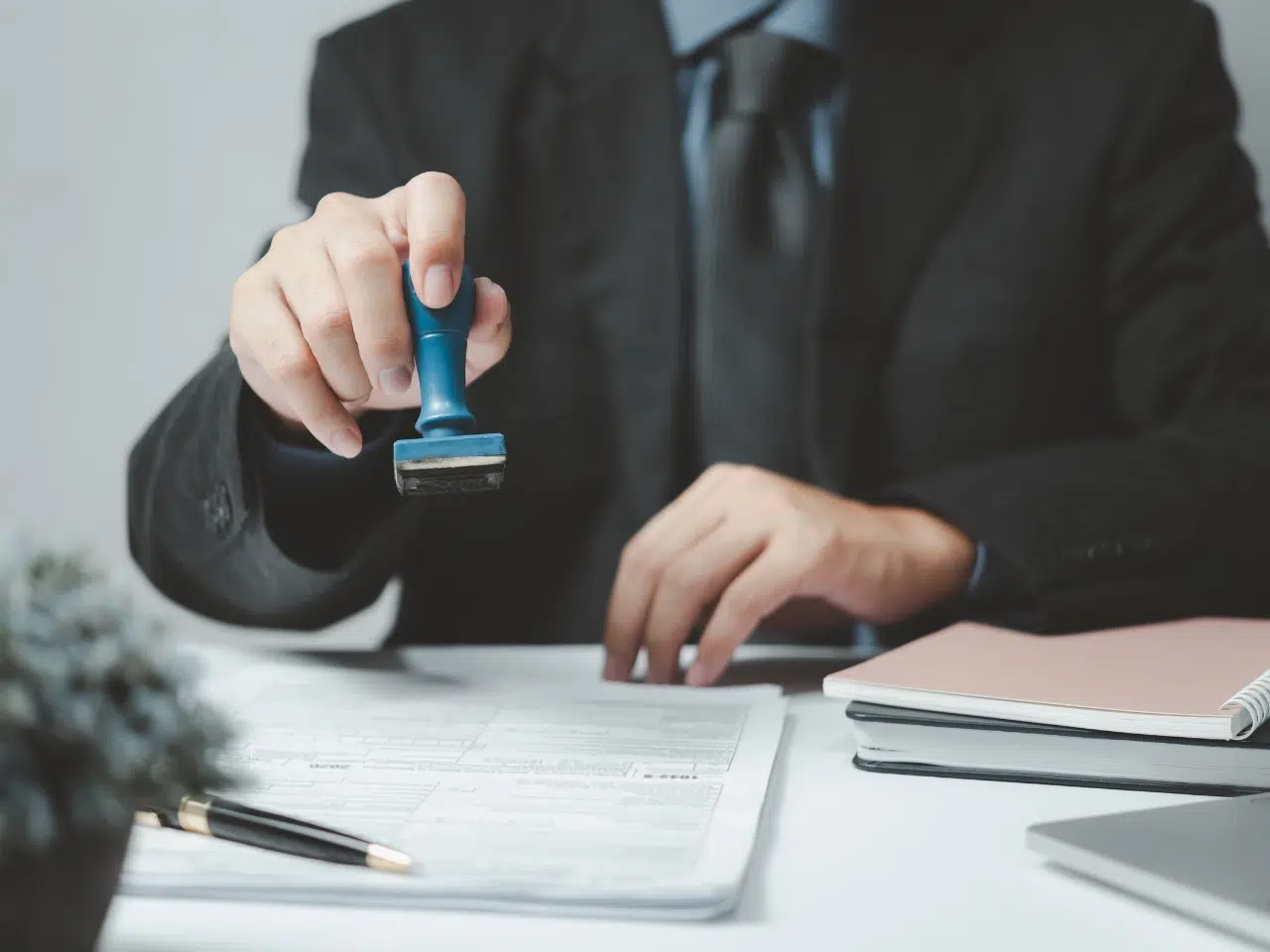 A person in a suit stamps a document on a desk, with a pen, papers, and a closed notebook nearby.