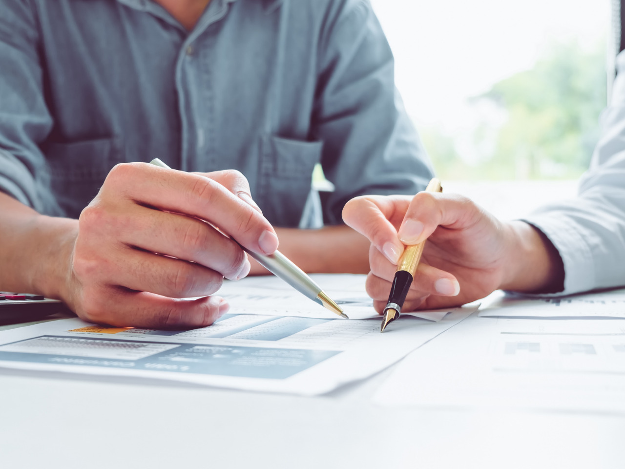 Two people sit at a table, discussing documents and charts while holding pens, focusing on paperwork, possibly collaborating on a business or financial project.