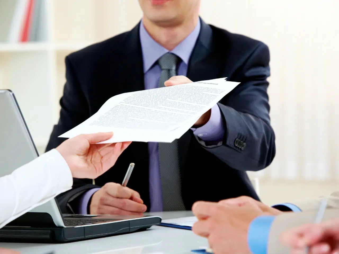 A person in a suit hands a document across a desk to another individual, with a laptop and another person’s hands visible in the foreground, suggesting a business meeting or contract signing.