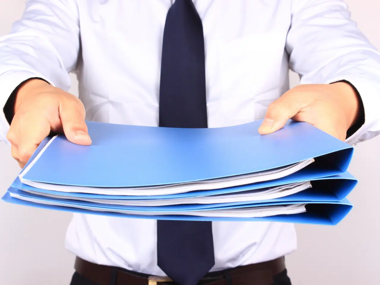 A person in a white shirt and black tie is holding out a stack of blue folders filled with papers, extending them towards the camera.