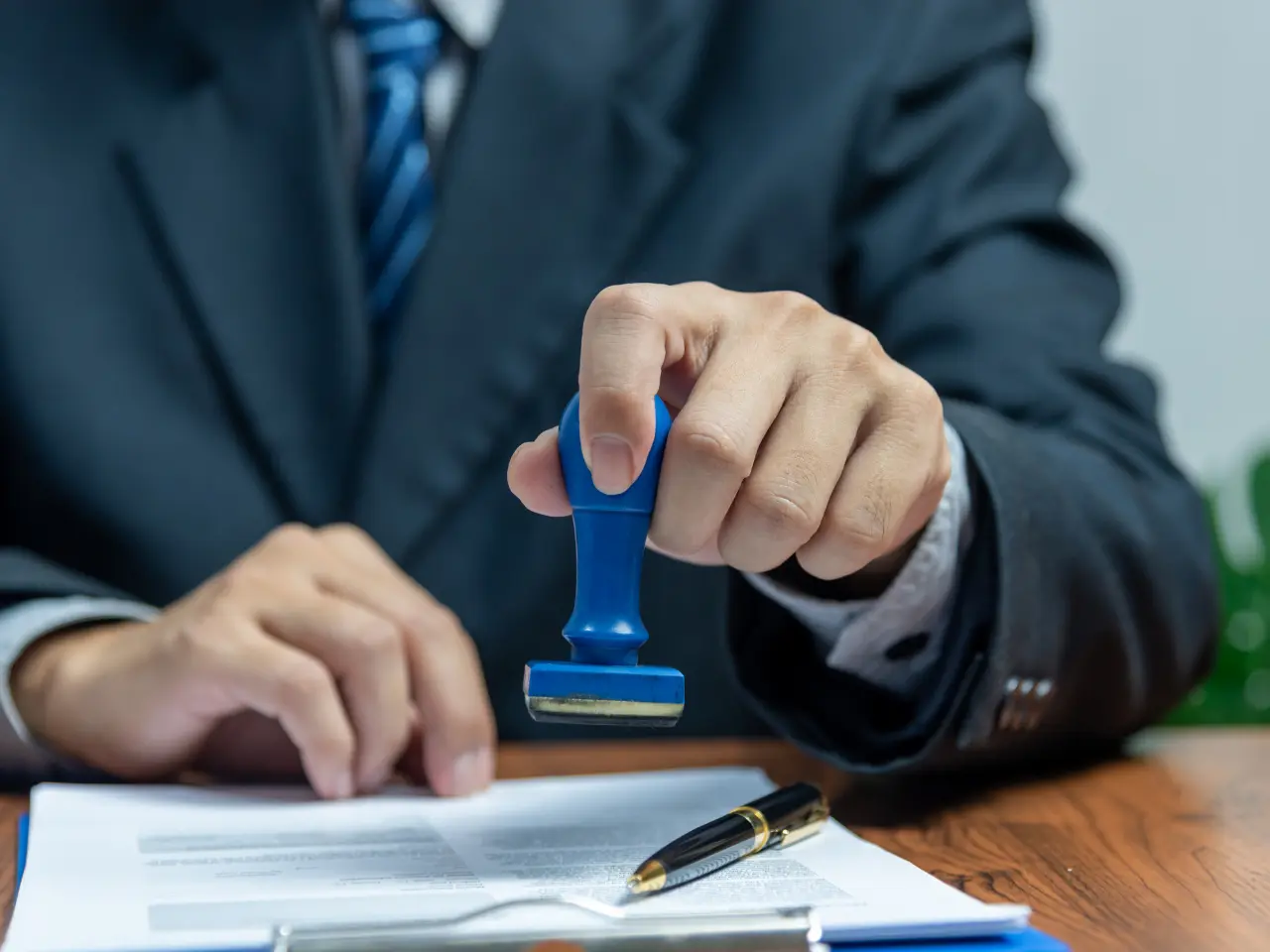 A person in a suit presses a blue stamp onto a document on a desk, with a pen and clipboard also visible.