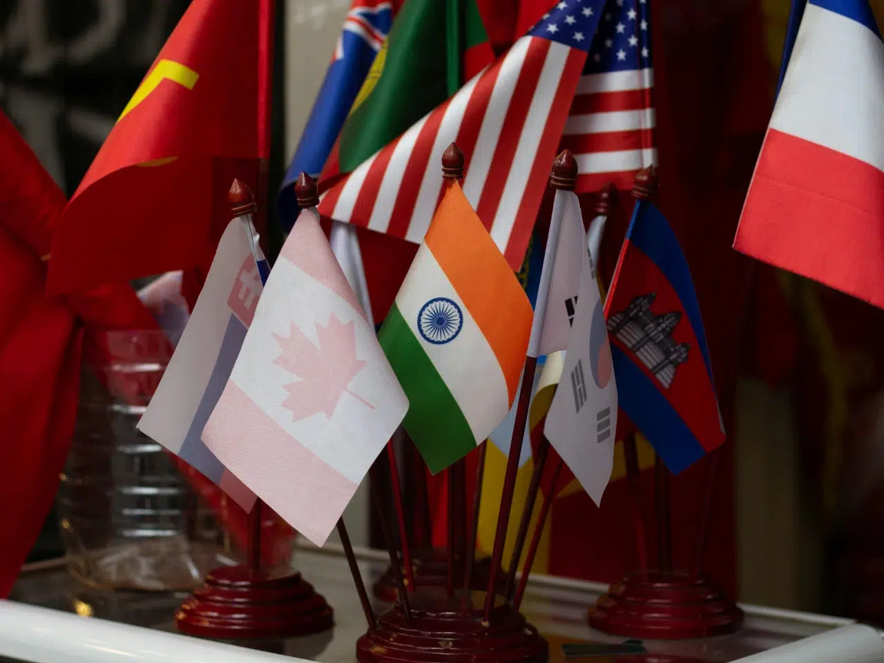 A group of small national flags, including Canada, India, the USA, South Korea, and others, displayed together on a table with red bases holding each flag upright.