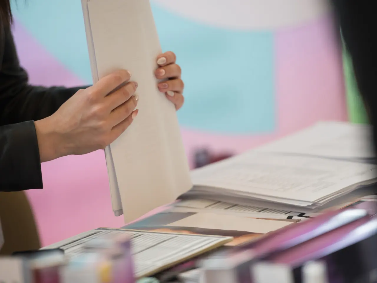 A person holding a folder and flipping through documents on a cluttered desk with papers, files, and a pastel-colored background.
