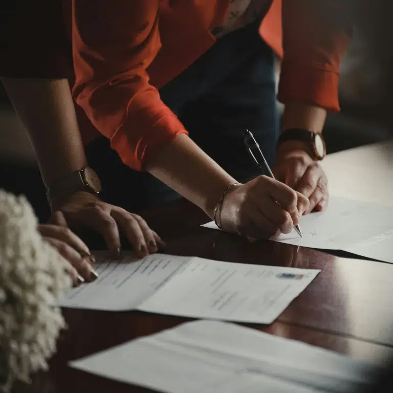 A person in a red jacket signs a document on a wooden table, while another person’s hand rests nearby on additional paperwork. The scene suggests a formal or official signing process.