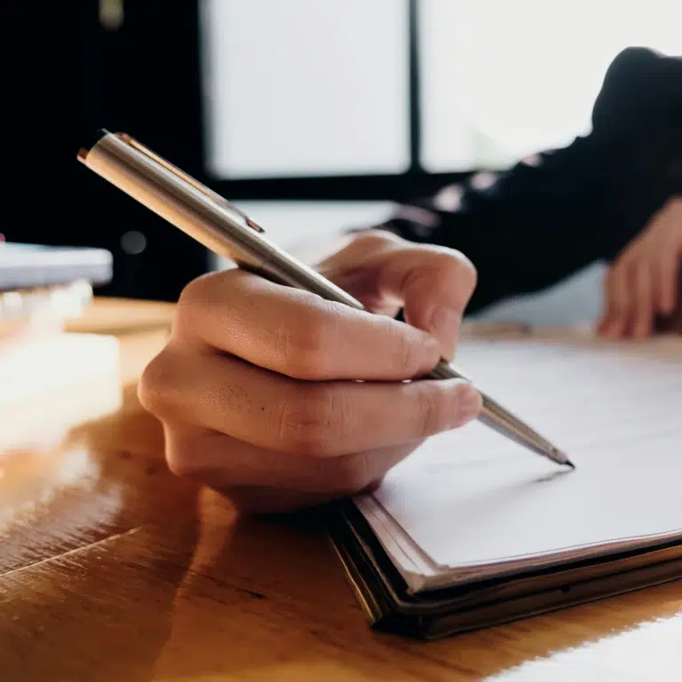 A close-up of a hand holding a pen and writing on a notepad on a wooden table, with sunlight coming through a window in the background.