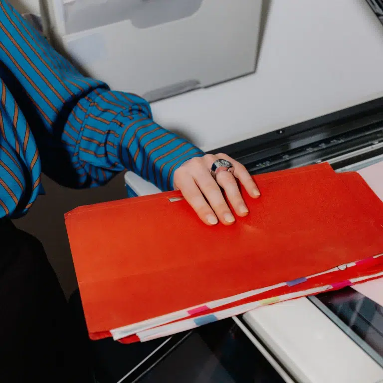 A person wearing a blue striped shirt holds a red folder over a photocopier, preparing to copy documents. Only the hand and part of the arm are visible.