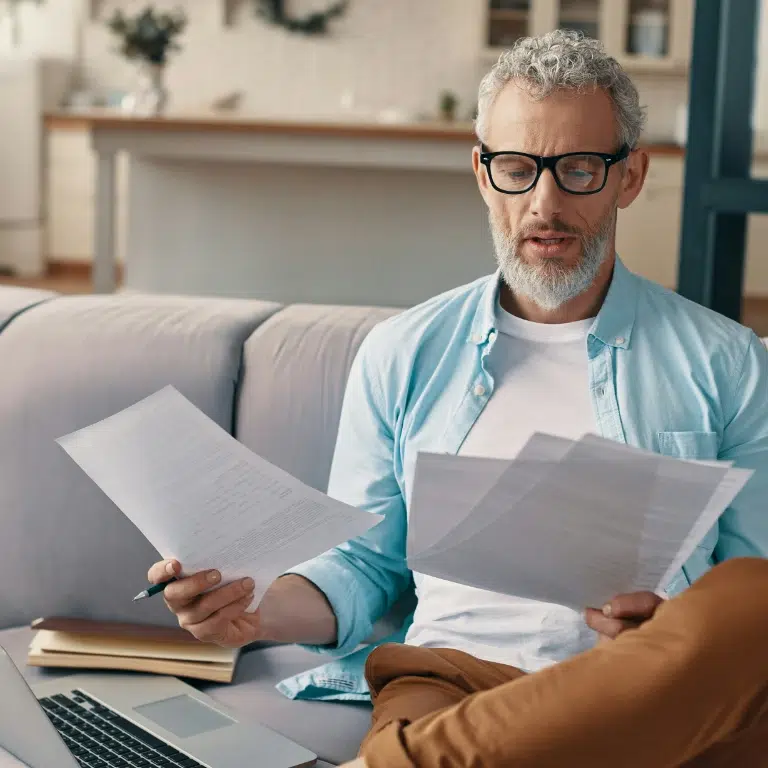 Middle-aged man with gray hair and glasses sitting on a couch, holding and reading documents, with a laptop, notebook, and pen beside him in a bright living room.