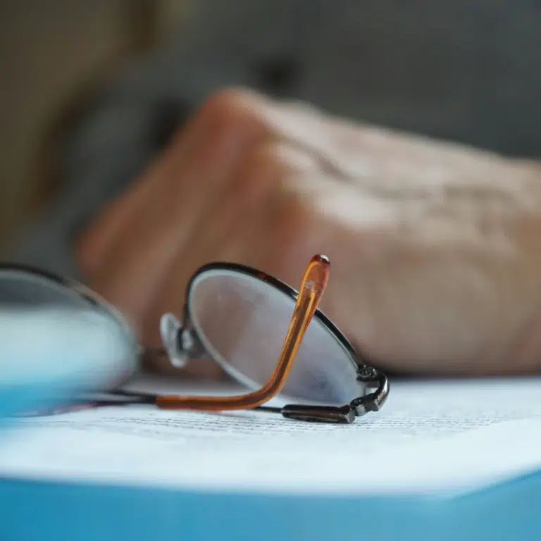 A close-up of eyeglasses resting on a document, with a persons blurred hand holding a pen in the background, suggesting reading or writing activity.
