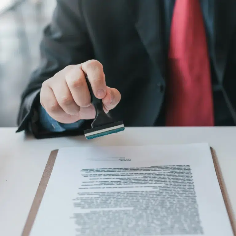 A person in a suit and red tie holds a rubber stamp above a document on a clipboard, suggesting approval or authorization of the paperwork.
