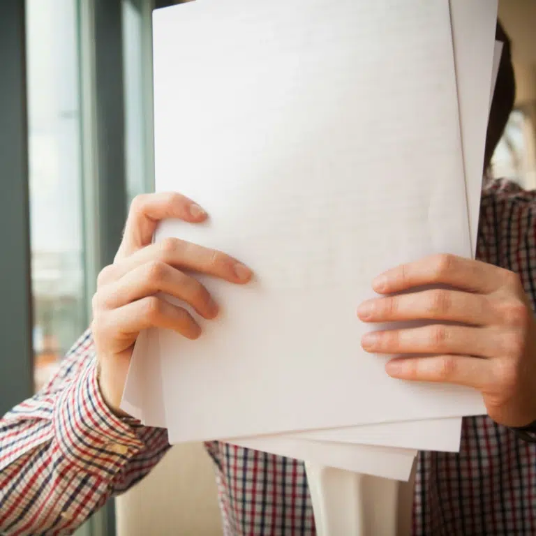 Two people are leaning over a table, signing documents. One person is wearing a watch and a red jacket, holding a pen, while the other persons hand rests on the papers. A bouquet of white flowers is visible in the foreground.