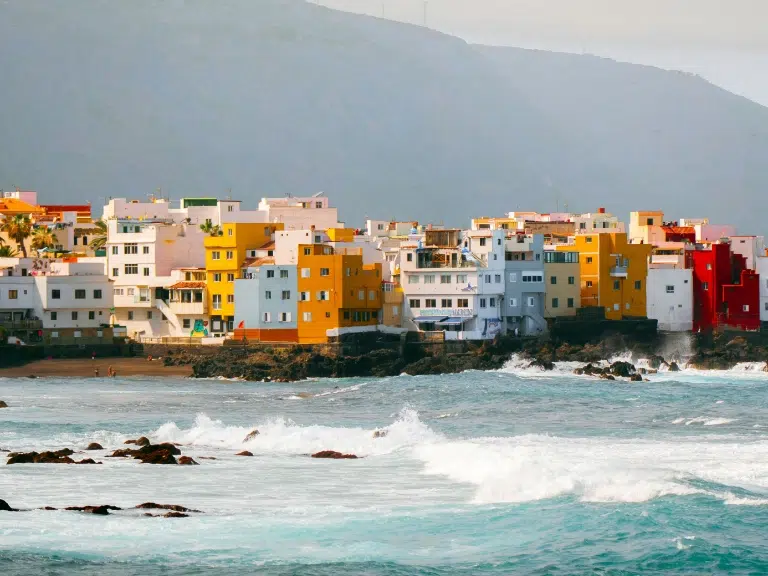 Colorful buildings line the rocky coastline with waves crashing against the shore, backed by a misty, mountainous landscape.