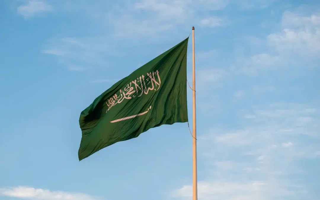 A green flag with white Arabic script and a sword, representing Saudi Arabia, waves on a flagpole against a blue sky with some light clouds.