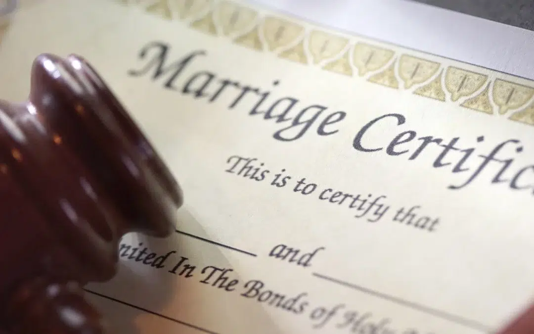 A close-up of part of a marriage certificate document with a wooden judge’s gavel resting on it. The text Marriage Certificate and some fill-in-the-blank lines are visible.