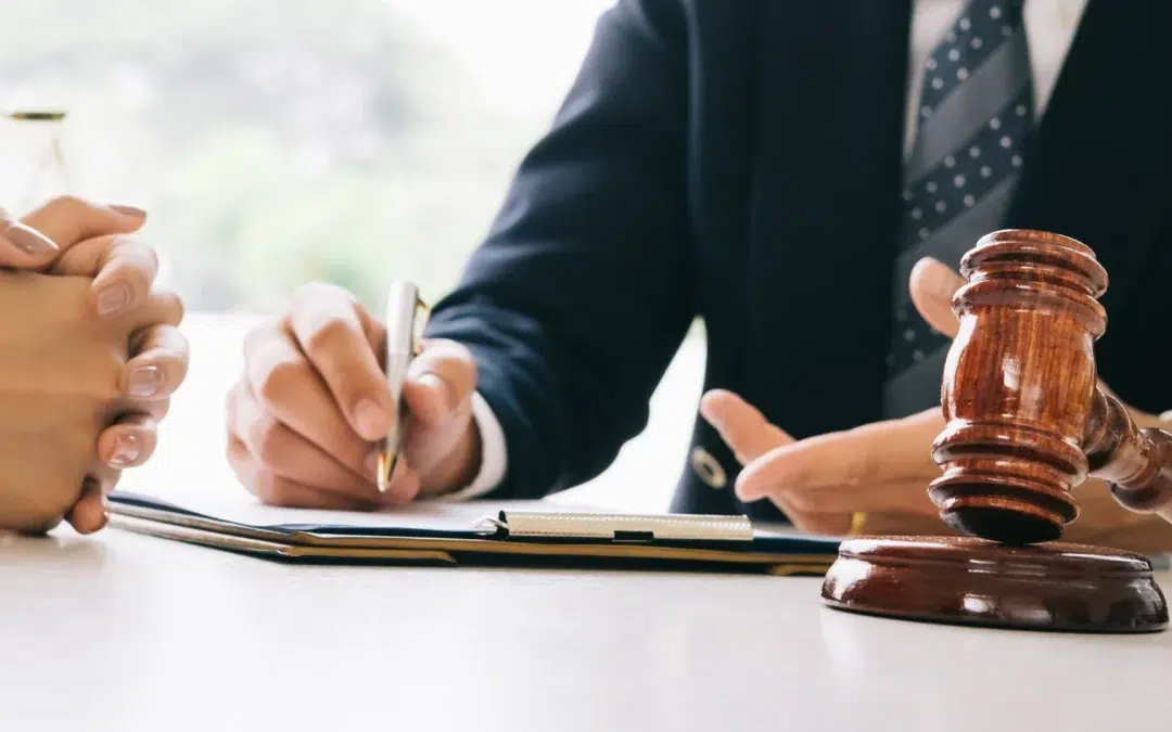 A person in a suit holds a pen and gestures while discussing documents with another person at a desk. A wooden judges gavel rests on the table in the foreground.