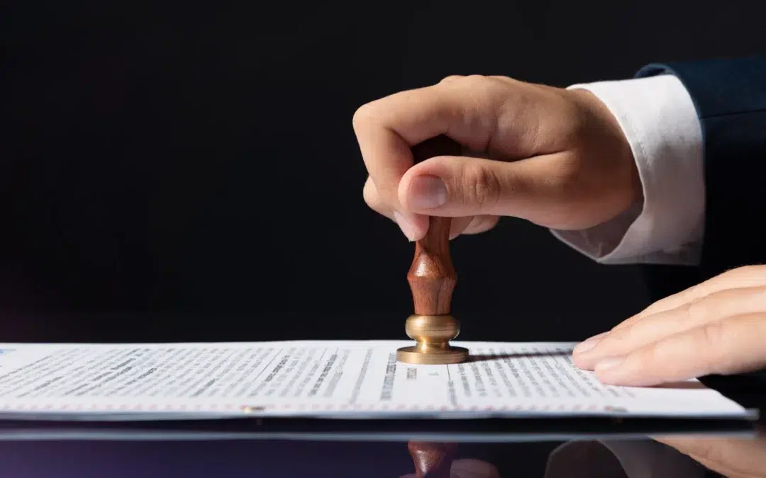 A person presses a wooden rubber stamp onto a document on a table, with the text on the page partially visible. The background is black, highlighting the action.
