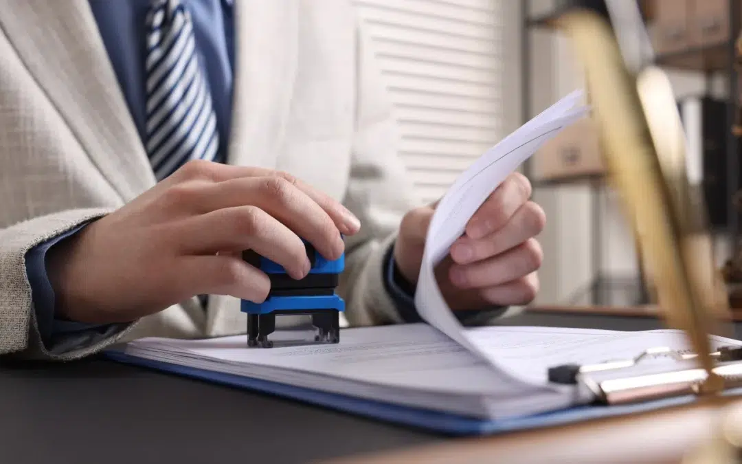 A person in a light-colored suit and striped tie stamps a document on a desk while holding a stack of papers. A clipboard, pen, and office items are visible in the background.