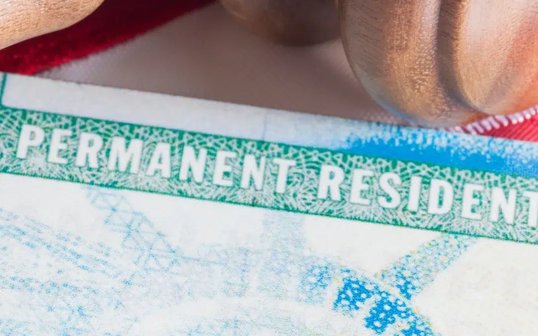 Close-up of the top portion of a U.S. Permanent Resident Card showing the words PERMANENT RESIDENT in white letters on a green background, with part of a flag and a wooden object visible in the background.