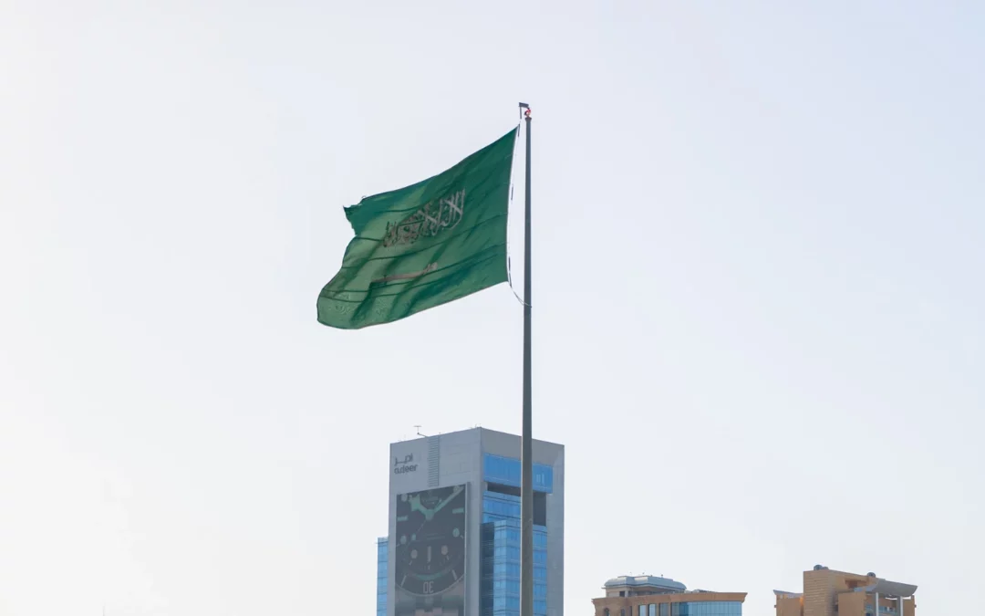 A green Saudi Arabian flag with white Arabic script and a sword flutters on a tall pole against a clear sky, with modern buildings visible in the background.