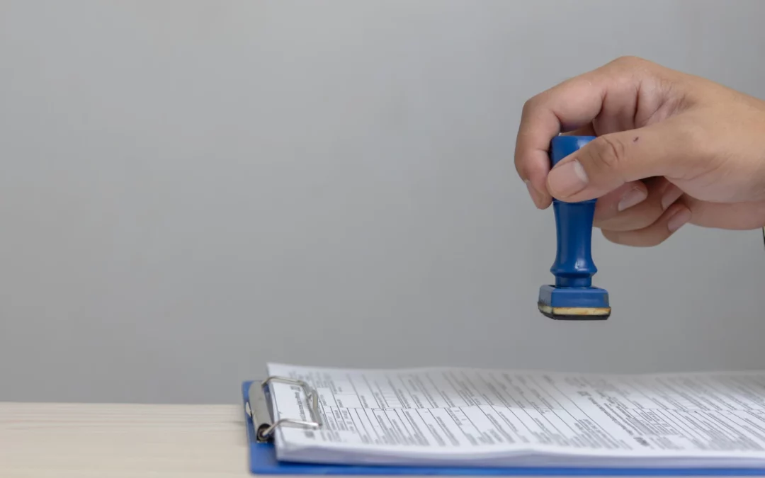 A hand holding a blue rubber stamp is about to stamp a document on a clipboard, which is placed on a light wooden desk against a plain gray background.