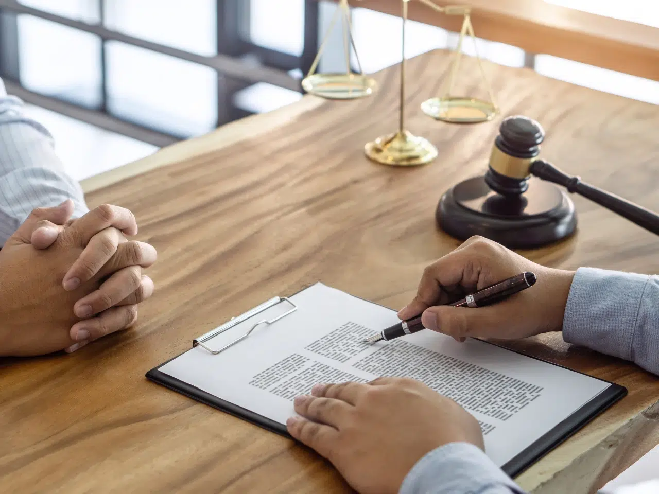 Two people sit across a wooden desk. One person points a pen at a contract on a clipboard while the other’s hands are clasped. A gavel and balance scales are on the desk, indicating a legal or consultation setting.