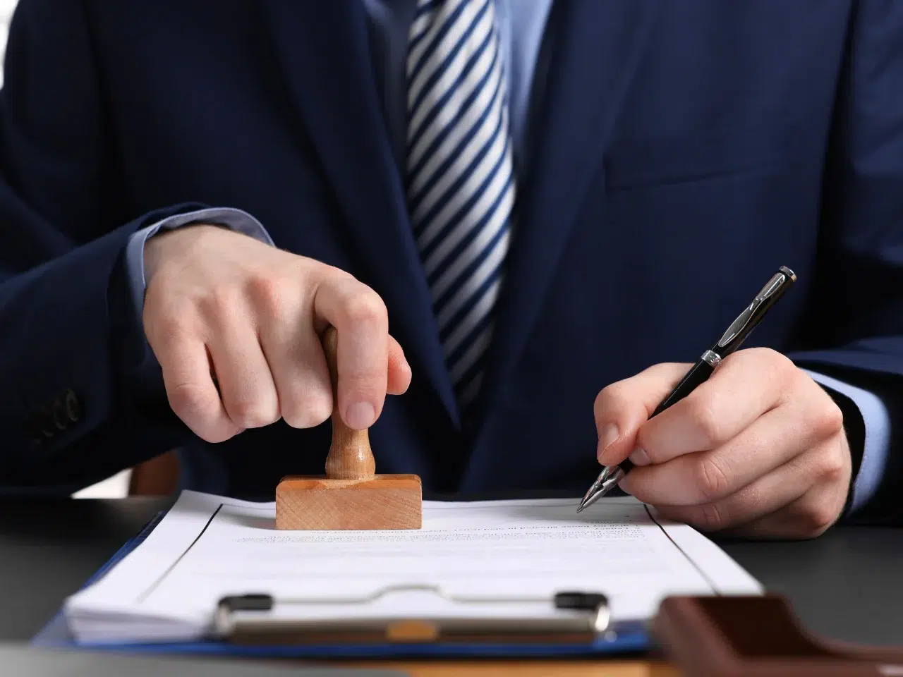 A person in a suit stamps a document with one hand and holds a pen in the other, sitting at a desk with paperwork.