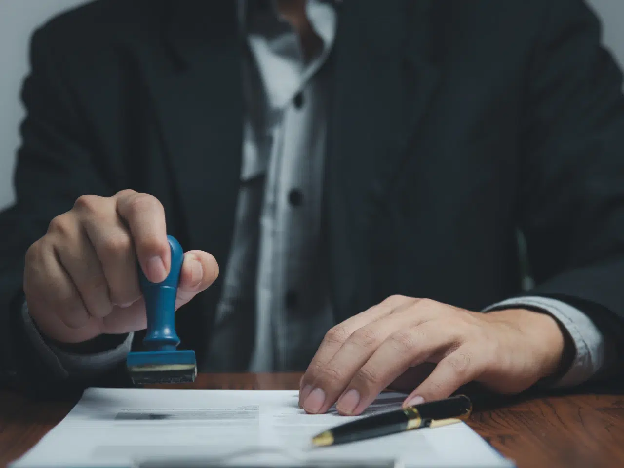A person in a suit uses a blue-handled stamp on a document at a desk, with a pen and papers nearby.