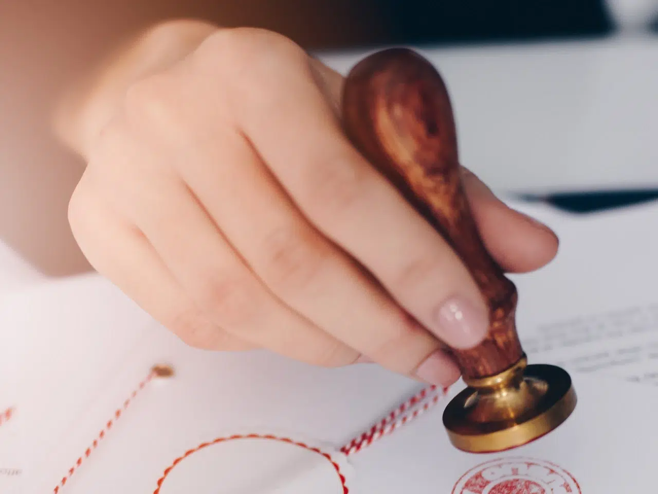 A close-up of a hand holding a wooden-handled stamp, pressing it onto a document with a red seal and string, indicating official approval or certification.