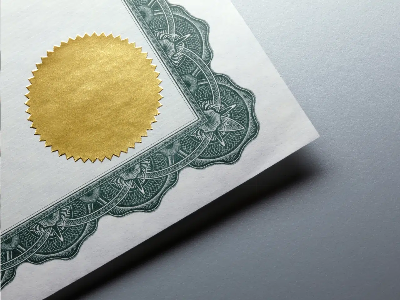 Close-up of a certificate corner featuring an ornate green border and a gold embossed seal on textured paper, set against a light gray background.