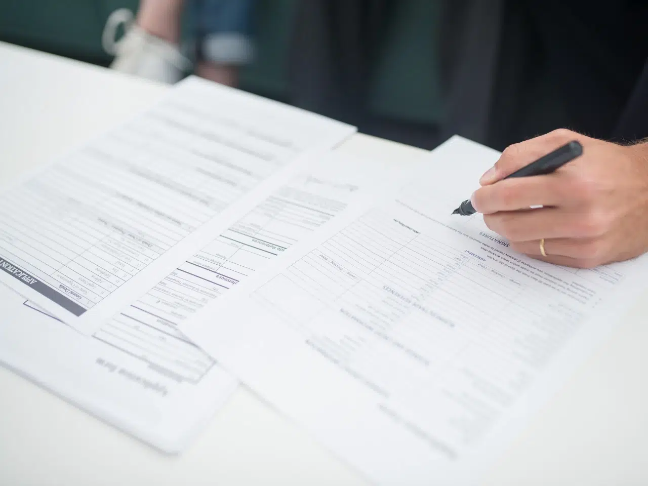 A hand holding a pen fills out a form on a desk, with additional documents and printed tables spread out nearby.