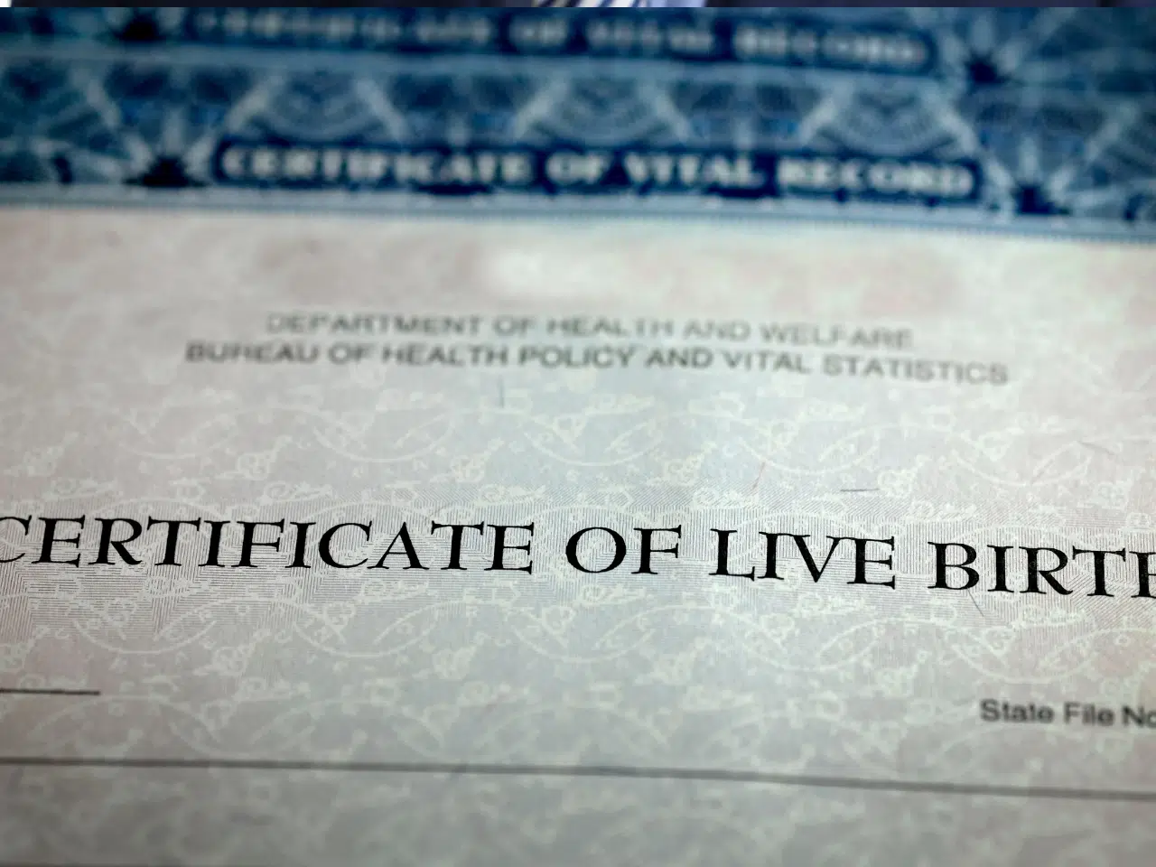 A close-up of a Certificate of Live Birth document with headings from the Department of Health and Welfare partially visible, showing official text and decorative border.