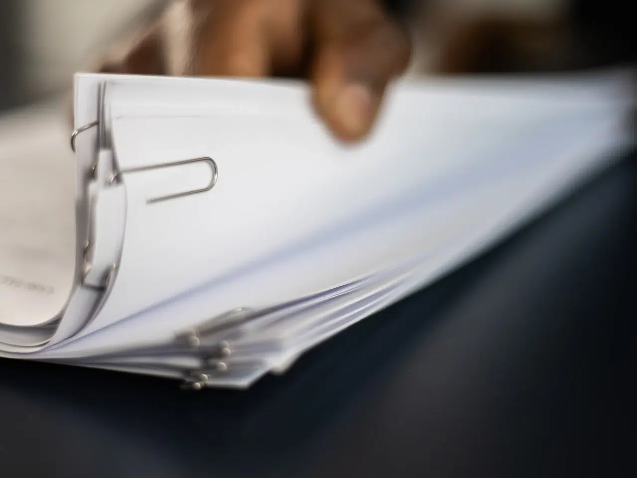 A close-up of a hand holding a stack of white papers secured with a large paperclip, resting on a dark surface.