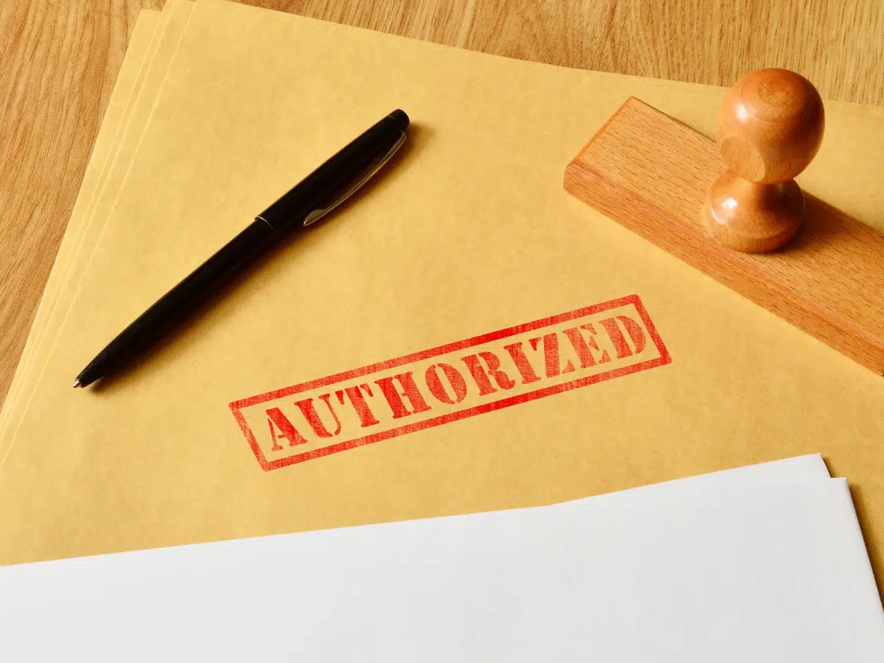 A black pen and a wooden stamp rest on a brown folder stamped with the word AUTHORIZED in red, next to a stack of white papers on a wooden desk.